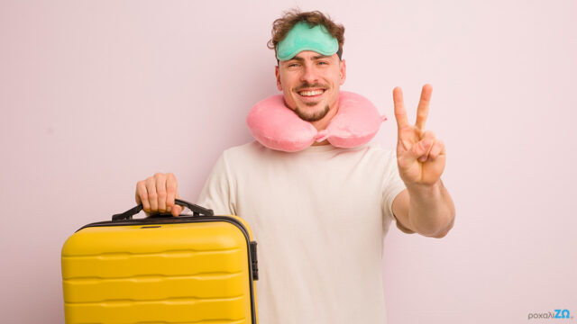 Sleep & Travel_Article Η εικόνα δείχνει έναν κύριο να κρατάει μία κίτρινη βαλίτσα , να φοράει μία μάσκα ύπνου κι ένα μαξιλαράκι αυχένα