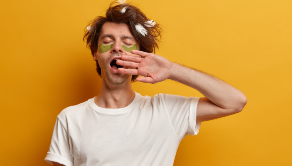 Ένας άντρας σε κίτρινο φόντο βάζει την παλάμη του μπροστά στο στόμα του και χασμουριέται.